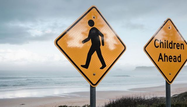 Coastal pedestrian crossing signs warning drivers near wet beach dunes with children ahead