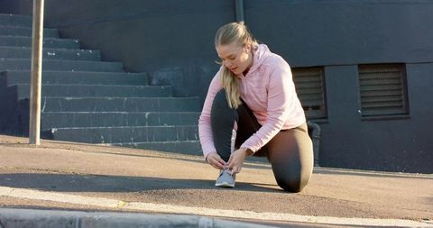 Young woman kneeling tying running shoe on urban sidewalk wearing pink hoodie and leggings