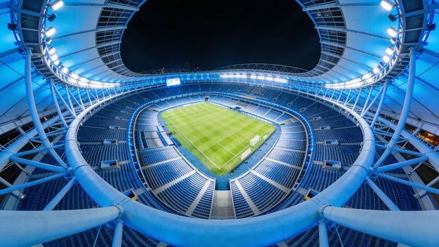 Aerial night view of modern floodlit stadium revealing blue seats and pristine football pitch