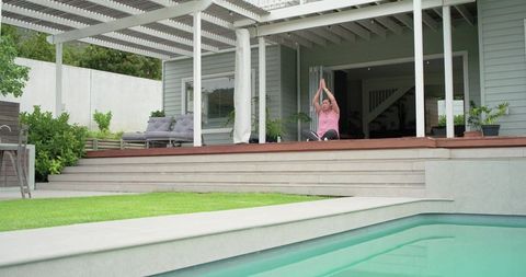 Mature woman practicing yoga on pergola deck overlooking pool and lawn