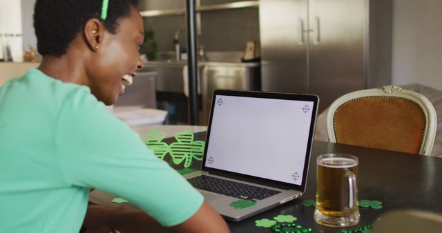 Woman is celebrating St. Patrick's Day indoors with green shamrock decorations. She is sitting in front of a laptop and laughing while enjoying a glass of beer. Ideal usage for holiday promotions, St. Patrick's Day marketing campaigns, and social media content highlighting festive gatherings.