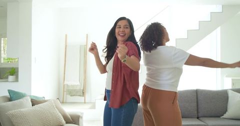 Multicultural women dancing and laughing in bright modern living room with natural daylight