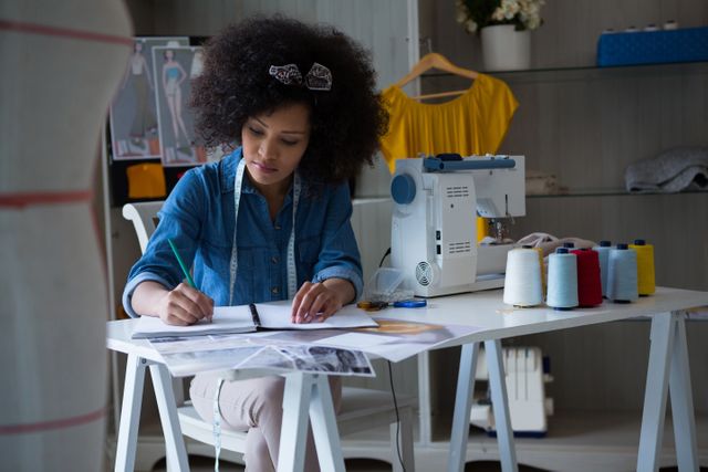 Female fashion designer sketching designs at her desk in a home studio. Sewing machine and spools of thread on the desk. Ideal for content related to fashion design, creativity, home-based businesses, and entrepreneurship.