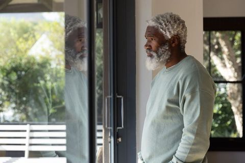 African american man standing by sliding glass door reflecting, looking contemplative in sunlit home