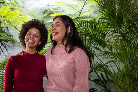 Diverse women laughing together in lush greenhouse with ferns and cozy sweaters