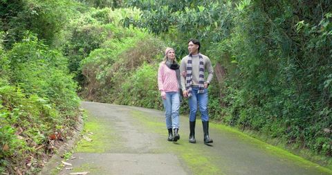 Couple walking mossy country lane in cozy sweaters, jeans and rubber boots under forest canopy