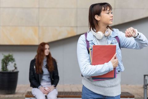 Young female student checking watch while holding notebooks with classmate on campus bench