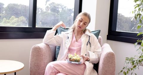 Female clinician in scrubs taking lunch break stretching neck while holding salad box