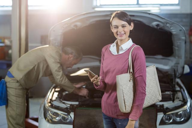 Portrait of customer holding a mobile phone while mechanic examining a car at the repair shop