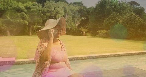 Senior woman wearing sunhat and sunglasses relaxing at poolside with floral wrap