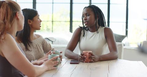Diverse women chatting over coffee around wooden table, woman with braids holding mug