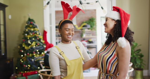 Two friends enjoying a Christmas celebration at home, one wearing a Santa hat and the other with reindeer antlers. They are standing near a decorated Christmas tree, smiling and sharing joy. The image is perfect for holiday-themed campaigns, festive social media posts, and marketing materials related to Christmas and togetherness.