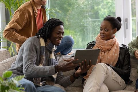 Friends sharing tablet and laughing on cozy sofa wearing scarves and jackets in daylight