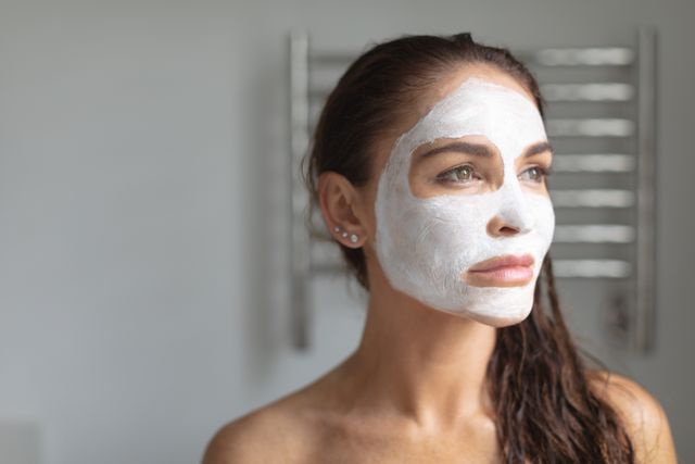 Thoughtful woman with facial mask standing in bathroom at home