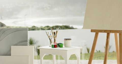 Sunlit artist workspace overlooking ocean featuring paintbrushes, paints and easel on patio