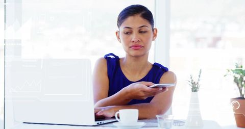 Woman holding smartphone and working on laptop while glancing left in bright coworking