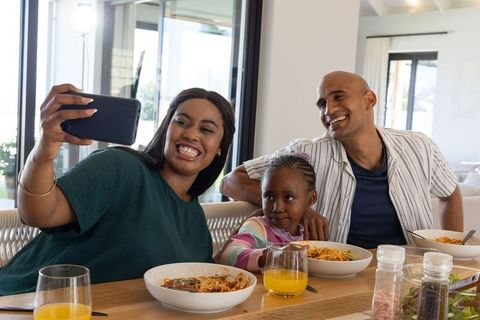 Multicultural family smiling and taking selfie while enjoying pasta at modern dining table
