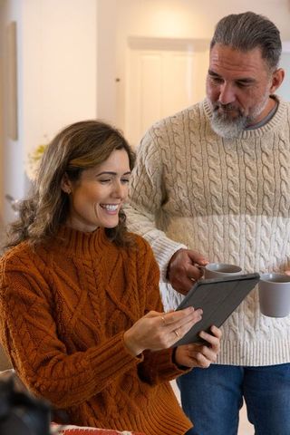 Middle-aged couple sharing coffee and browsing tablet in cozy cable-knit sweaters at home
