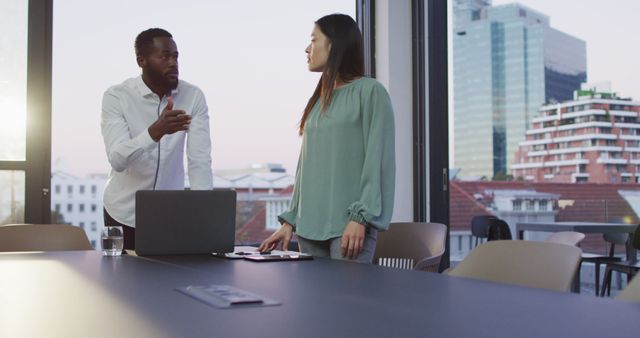 Businesspeople in a professional setting discussing work in a modern office with a city view. One person stands while the other sits, indicating an active discussion. Ideal for use in business blogs, corporate websites, advertisements for office spaces, presentations, and articles focused on teamwork and professional environments.