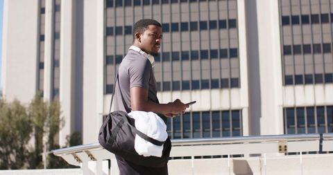 African American man standing on urban terrace checking smartphone wearing headphones carrying gym b