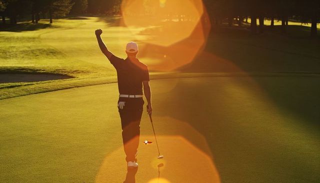 Walking golfer raising fist in victory on putting green at sunset with lens flare and golden hour li