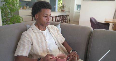 Young African American Man Working Remotely on Laptop While Holding Pink Mug on Gray Sofa