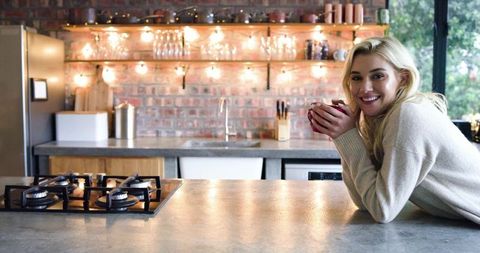 Smiling woman leaning on kitchen island holding mug in rustic modern kitchen