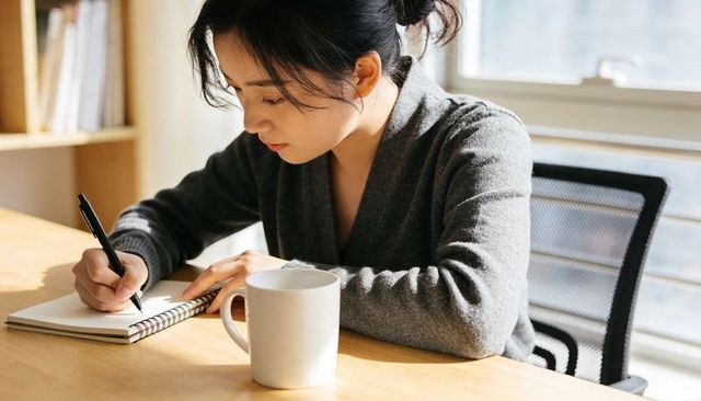 Focused woman writing in spiral notebook at sunlit desk with coffee, working from home