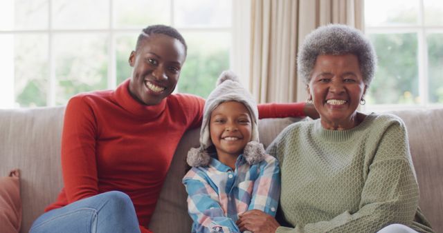 Showcasing joyful moments and strong family bonds, this image features three generations of African American females sitting together on a couch, smiling warmly. Ideal for use in family-related advertising, blog posts about family values, intergenerational activities, or promotional materials for family services.