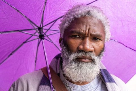 Thoughtful senior black man under purple umbrella on rainy urban street wearing scarf