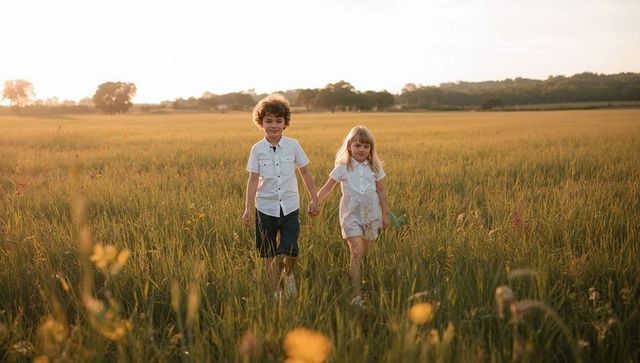 Siblings walking hand-in-hand through golden meadow at sunset, backlit childhood moment