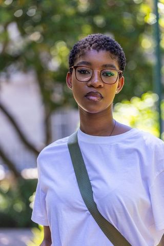 African woman standing in sunlit park wearing tortoiseshell glasses and green bag strap