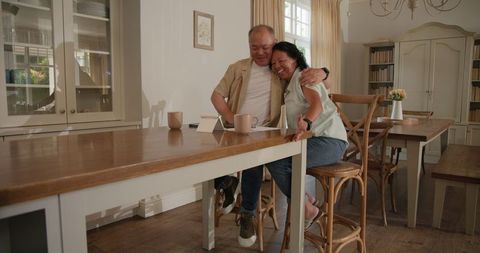 Happy Senior Asian Couple Using Tablet While Sitting in Warm Dining Room