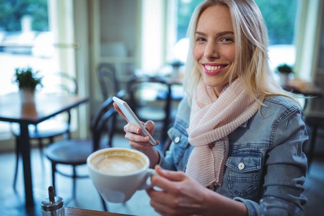 Woman enjoying coffee while using smartphone in a cozy café. Ideal for lifestyle blogs, technology advertisements, and social media content promoting relaxation and modern living.