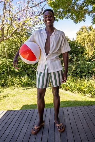 Smiling man holding colorful beach ball on sunny backyard deck in open shirt and striped shorts