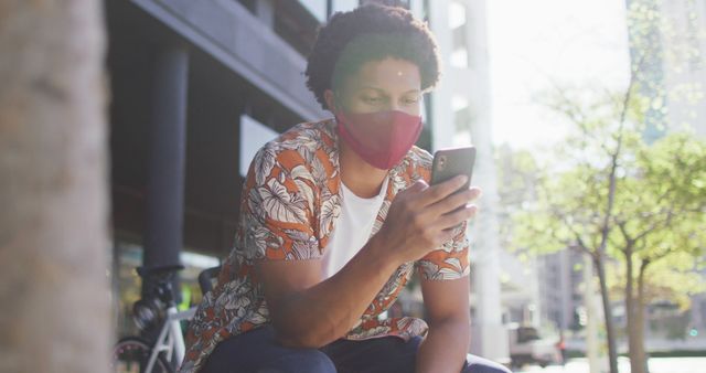 Man sitting outside in an urban environment, wearing floral shirt and face mask, engaged with smartphone. Could be used to depict new normal germain protocols, tech-savvy lifestyle, or city living dynamics.