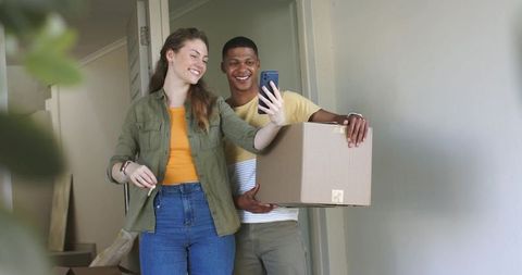 Smiling multicultural couple moving into new home taking selfie with smartphone and box