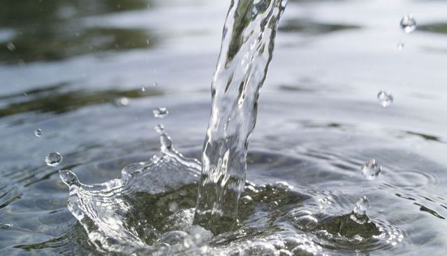Pouring water forming crown splash and concentric ripples on calm pool with soft reflections