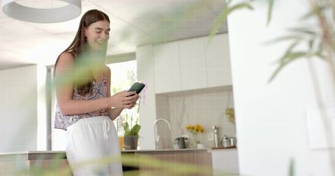 Smiling woman using smartphone in bright modern kitchen with yellow flowers