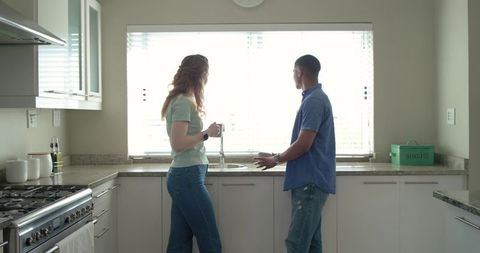 Diverse couple standing and looking out window at kitchen sink in sunlit modern kitchen