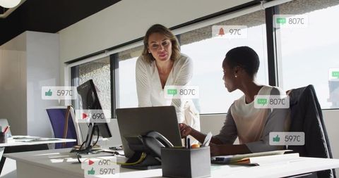 Businesswomen collaborating over laptop in bright modern office showing social metrics