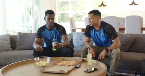 Diverse friends sitting on sofa sharing pizza and drinks in sunlit modern living room