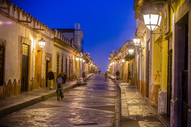 This image showcases a picturesque colonial street illuminated by warm vintage street lamps at night. Cobblestone paths stretch along. Few people are seen walking or riding bicycles, adding life to the serene atmosphere. Ideal for use in travel blogs, tourism promotions, history and architecture articles, postcards, or cultural documentaries to evoke feelings of nostalgia and romanticism.