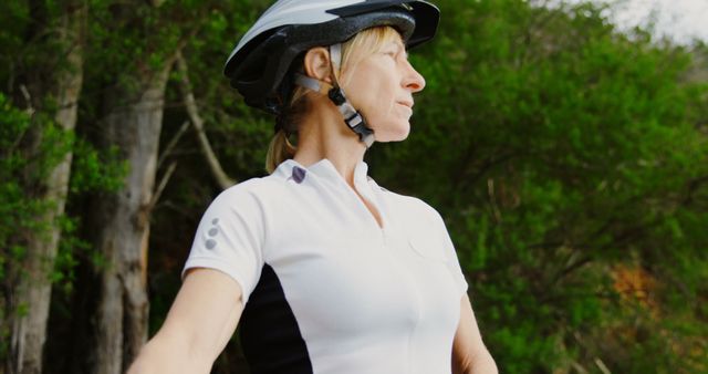 Mature woman cyclist wearing helmet posing outdoors in a nature setting with trees in the background. Ideal for promoting active lifestyles, senior fitness, cycling gear, and outdoor activities. Suitable for health and wellness articles, sports promotions, adventure magazines.