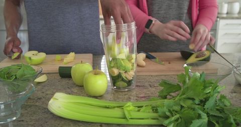 Couple preparing green smoothie chopping apples avocado celery filling clear blender cup
