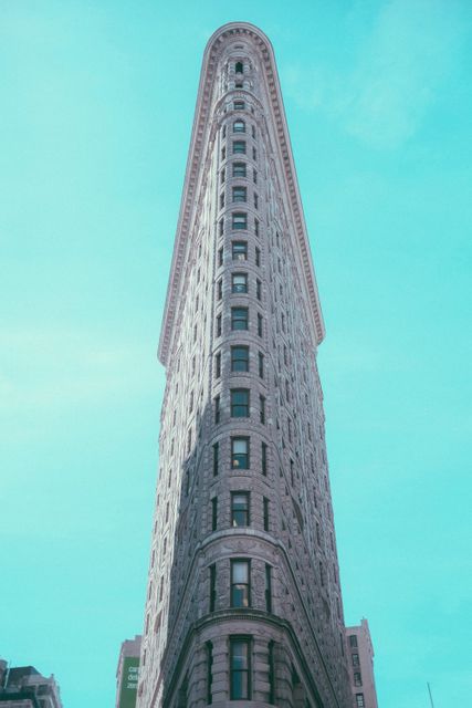 Image of the iconic Flatiron Building in New York City captured against a clear blue sky. The triangular shape and intricate architecture of the historic skyscraper are prominently featured. Ideal for use in travel articles, architecture magazines, urban exploration content, and promotional materials for New York City tours.