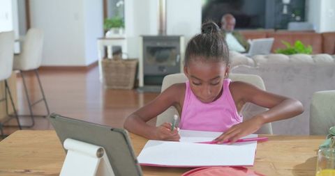 Young girl doing homework at tablet on wooden table in open-plan living room with parent
