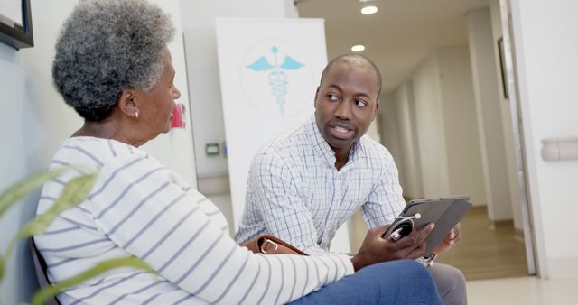 Male healthcare professional consulting senior African American female patient in a modern medical office. Both are engaged in a thoughtful conversation with a medical clipboard and device. Ideal for illustrating healthcare services, doctor-patient relationships, elderly care, and medical office teamwork.