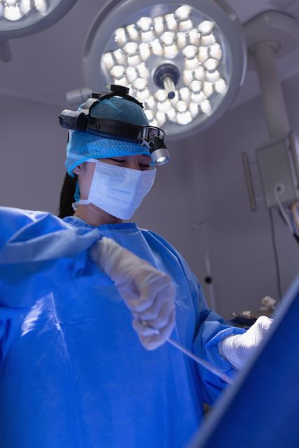 This image depicts a female surgeon in a hospital operating room, performing a surgical procedure. She is wearing surgical scrubs, a mask, and gloves, with surgical lights overhead. This image can be used for medical and healthcare-related content, including articles, brochures, and educational materials about surgery, healthcare professionals, and hospital environments.