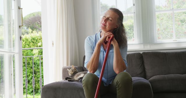 Middle-aged woman sitting on a cozy couch in a sunlit living room, staying close to her pet cat. Perfect for themes related to tranquility, home life, companionship, self-reflection, and cozy living spaces.